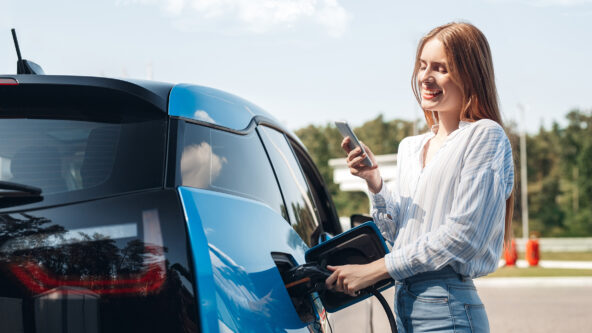 Young beautiful woman traveling by electric car having stop at charging station standing plugging cable browsing internet on smartphone smiling joyful while charing